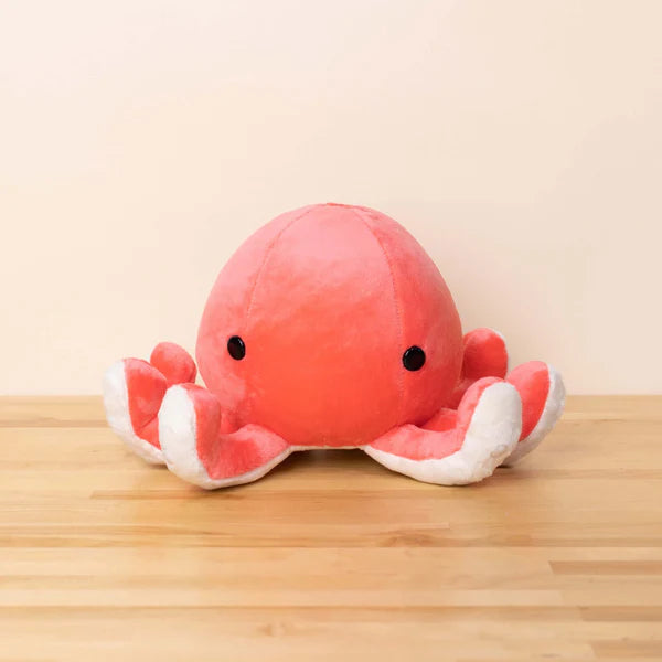 Red and white plush octopus toy on a wooden surface with a beige background. It has upturned tentacles and round black eyes.