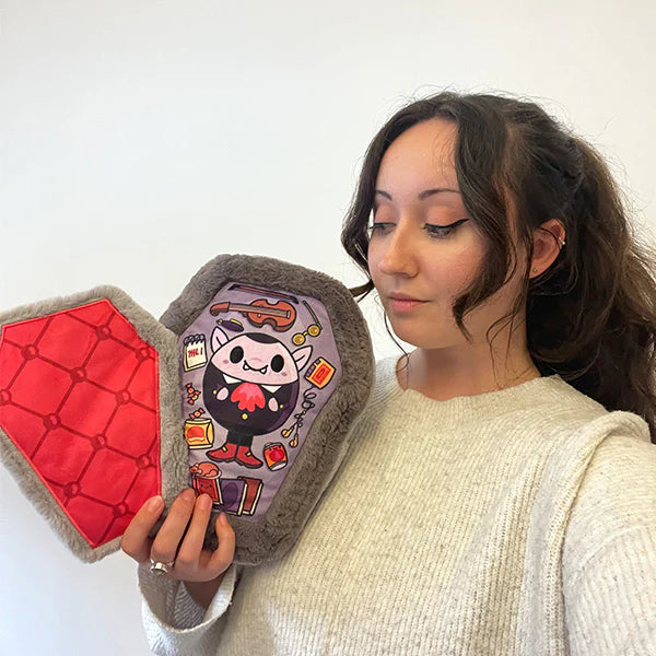 Woman holding the plush coffin with the lid open, showing the printed inner details, against a white background.