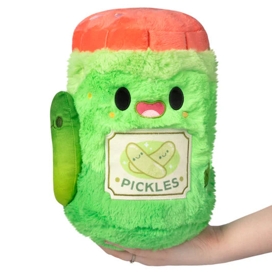 Green plush toy resembling a jar of pickles with a red lid, held in a hand against a white background.