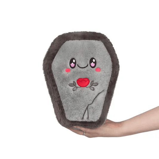 Plush toy resembling a coffin with a face and rose design, held by a hand on a white background. The plush coffin opens to reveal a printed inside with details of a vampire with books, a violin, snacks and more.