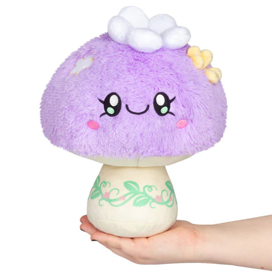 Plush toy mushroom with a smiling face held in a hand on a white background. The cap is purple with plush white and yellow flowers attached, and the stem is tan with a printed leaf design.