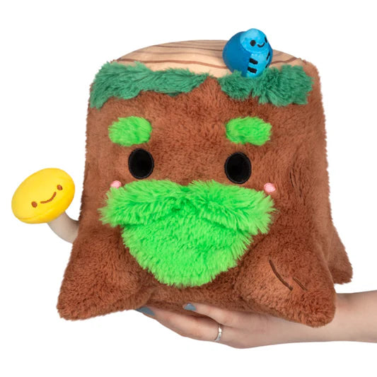 Plush toy resembling a tree stump with a face, held by a hand on a white background. The stump has additional plush details, such as fuzzy green eyebrows and a fuzzy green beard, a plush yellow mushroom attached, and a blue plush bug on top.