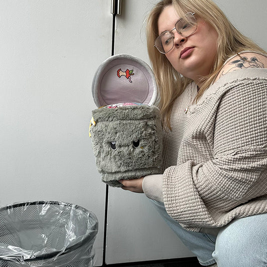Person holding a plush toy resembling a trash can in front of a trash can.