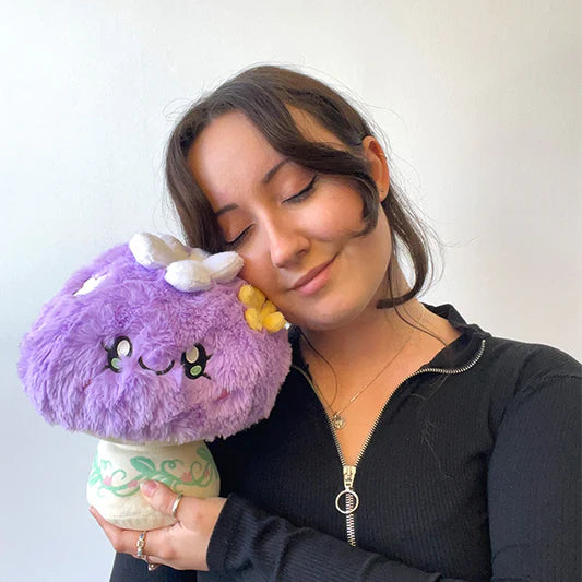 Person holding a purple plush toy mushroom with a white flower and a yellow flower attached and a smiling face on a plain background